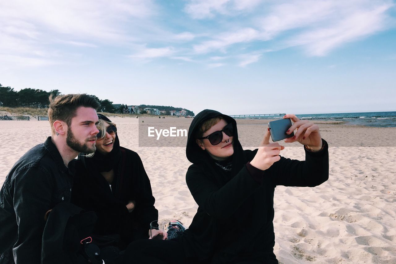 Young friends taking selfie at beach against sky