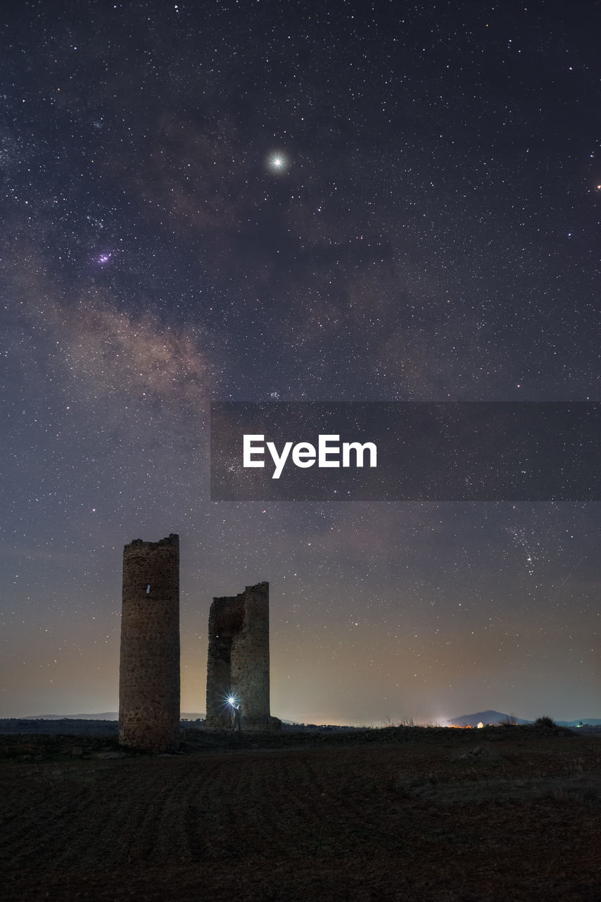 Anonymous person with lantern light on ancient stone towers on empty sandy ground under dark starry sky with milky way