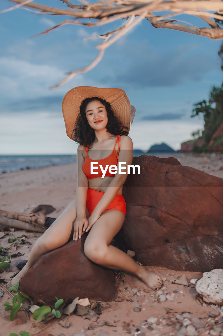 portrait of young woman sitting on rock at beach against sky