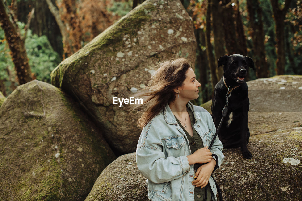 Girl looking at a black dog on a leash sitting on rock in the forest