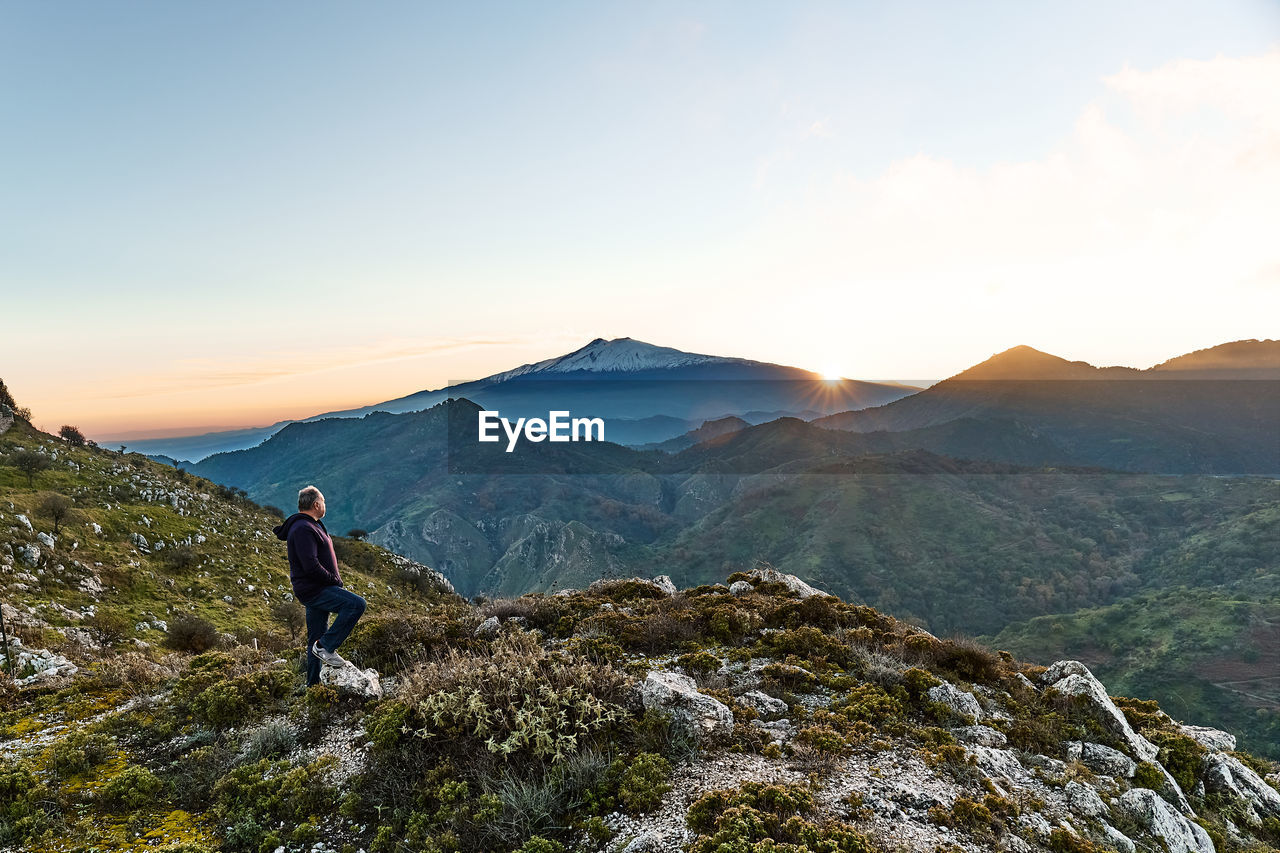 Man standing on top of the cliff in mountains at sunset enjoying sunset over erupting volcano etna.