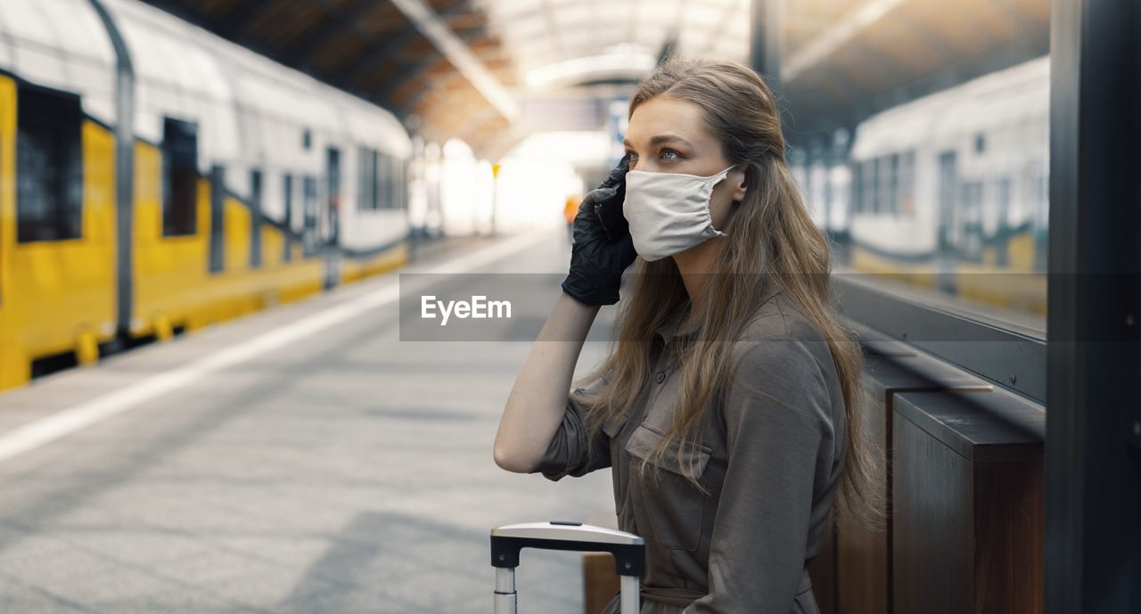 Side view of woman wearing mask talking over smart phone while sitting at railroad station