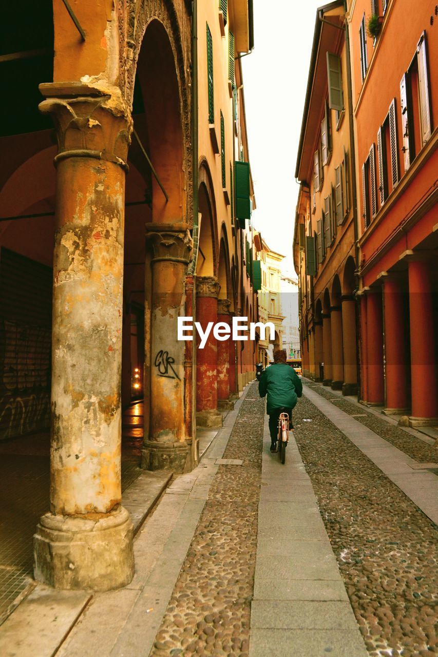 REAR VIEW OF MAN WALKING ON STREET AMIDST BUILDINGS