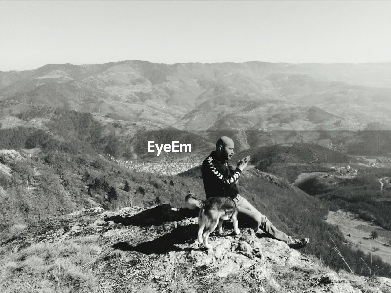 Side view of man sitting on cliff with dog against sky