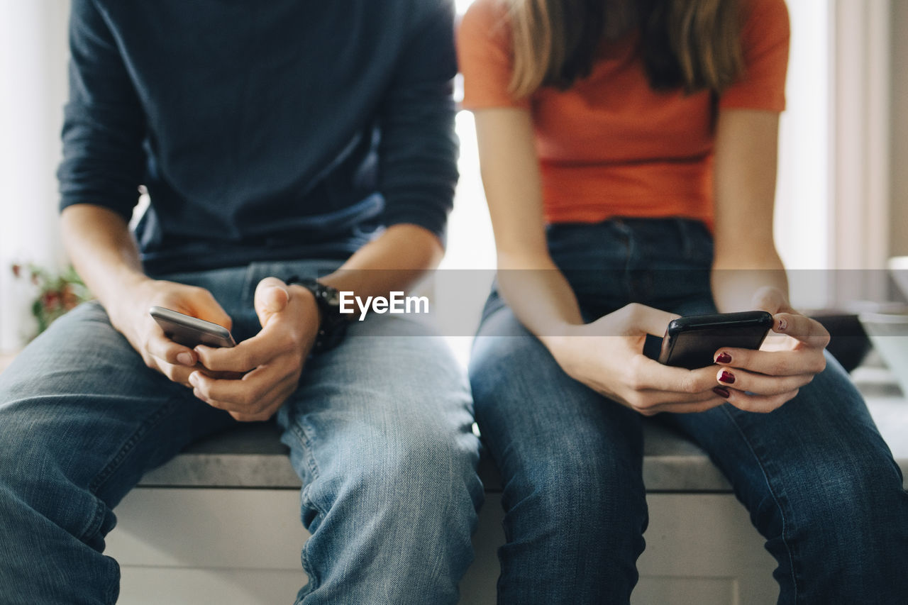 Midsection of male and female friends using mobile phones while sitting at kitchen counter