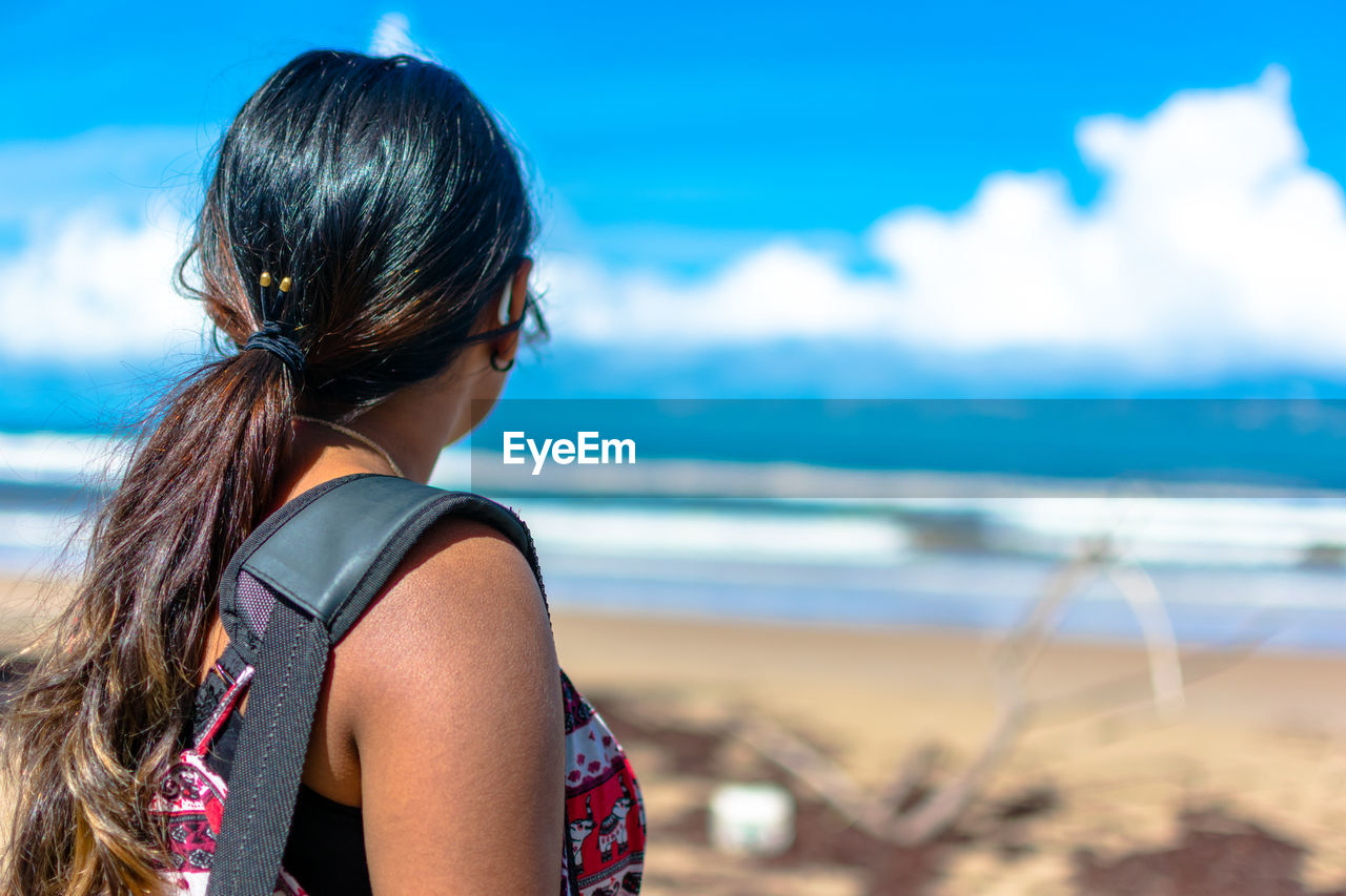 Side view of young woman with backpack standing at beach against blue sky