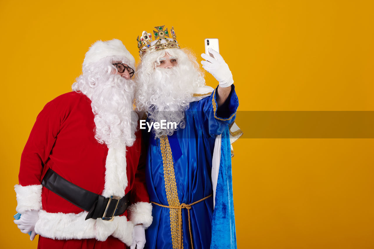 santa claus, colored background, celebration, adult, men, studio shot, costume, facial hair, beard, indoors, senior adult, dressing up, holiday, yellow, clothing, traditional clothing, waist up, yellow background, person, two people, happiness, portrait, emotion, christmas, seniors, smiling, fun, gray hair, standing, togetherness, humor