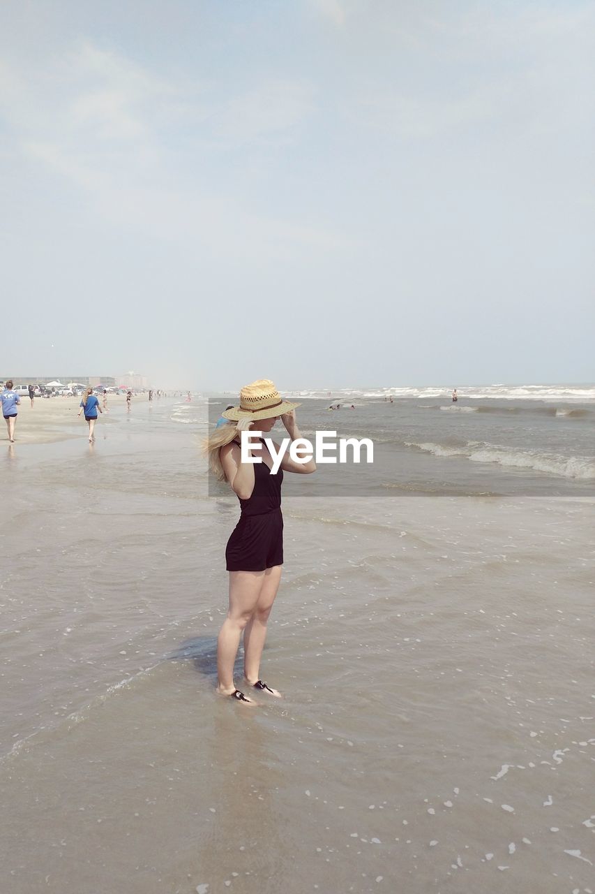 Full length of woman wearing hat standing on shore at beach