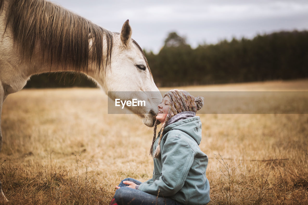 White horse tugging on girls hat in field in the fall while girl sits