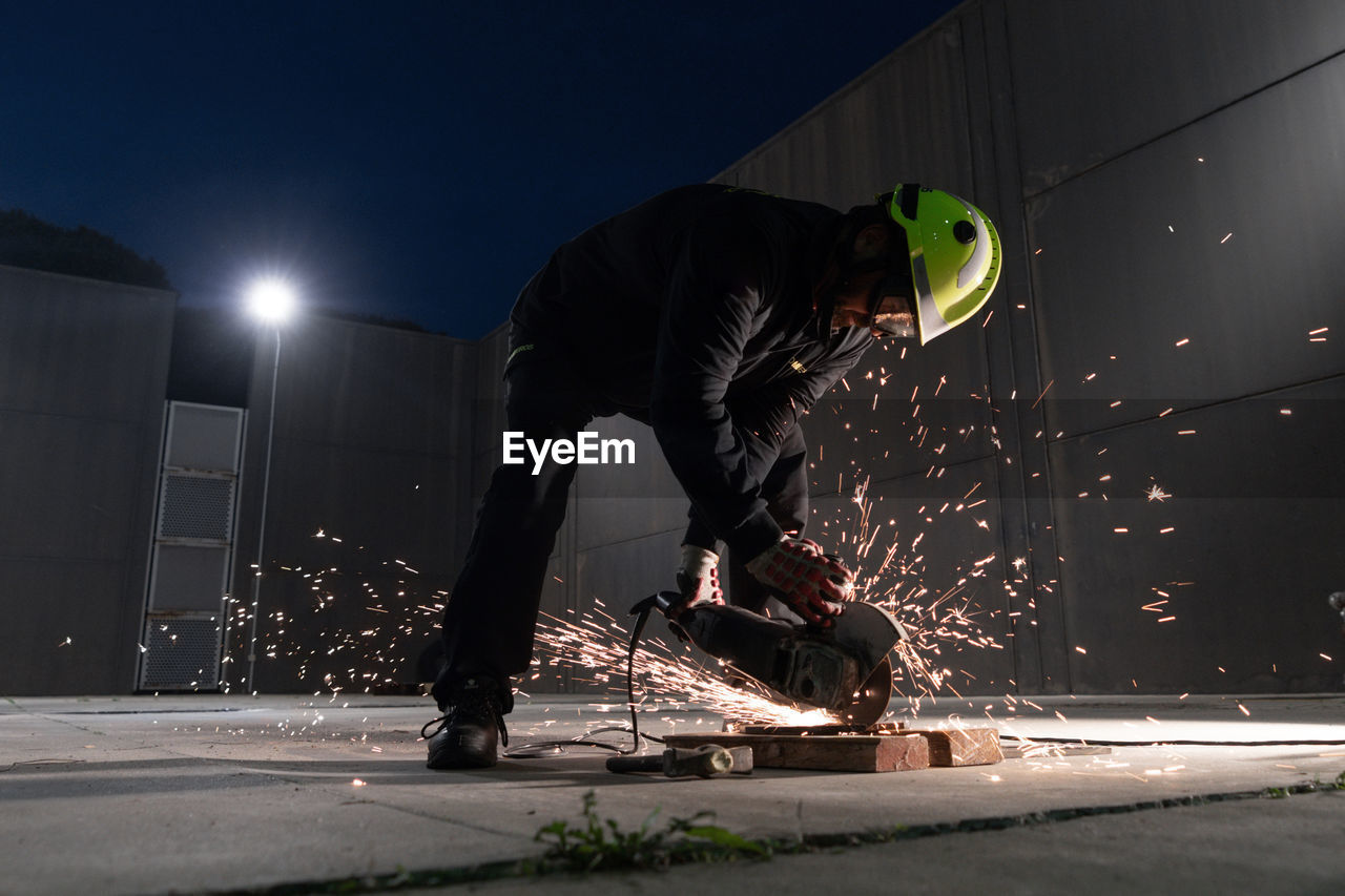 Full body of unrecognizable male welder in protective helmet and goggles cutting iron details with chop saw during work at industrial factory at night