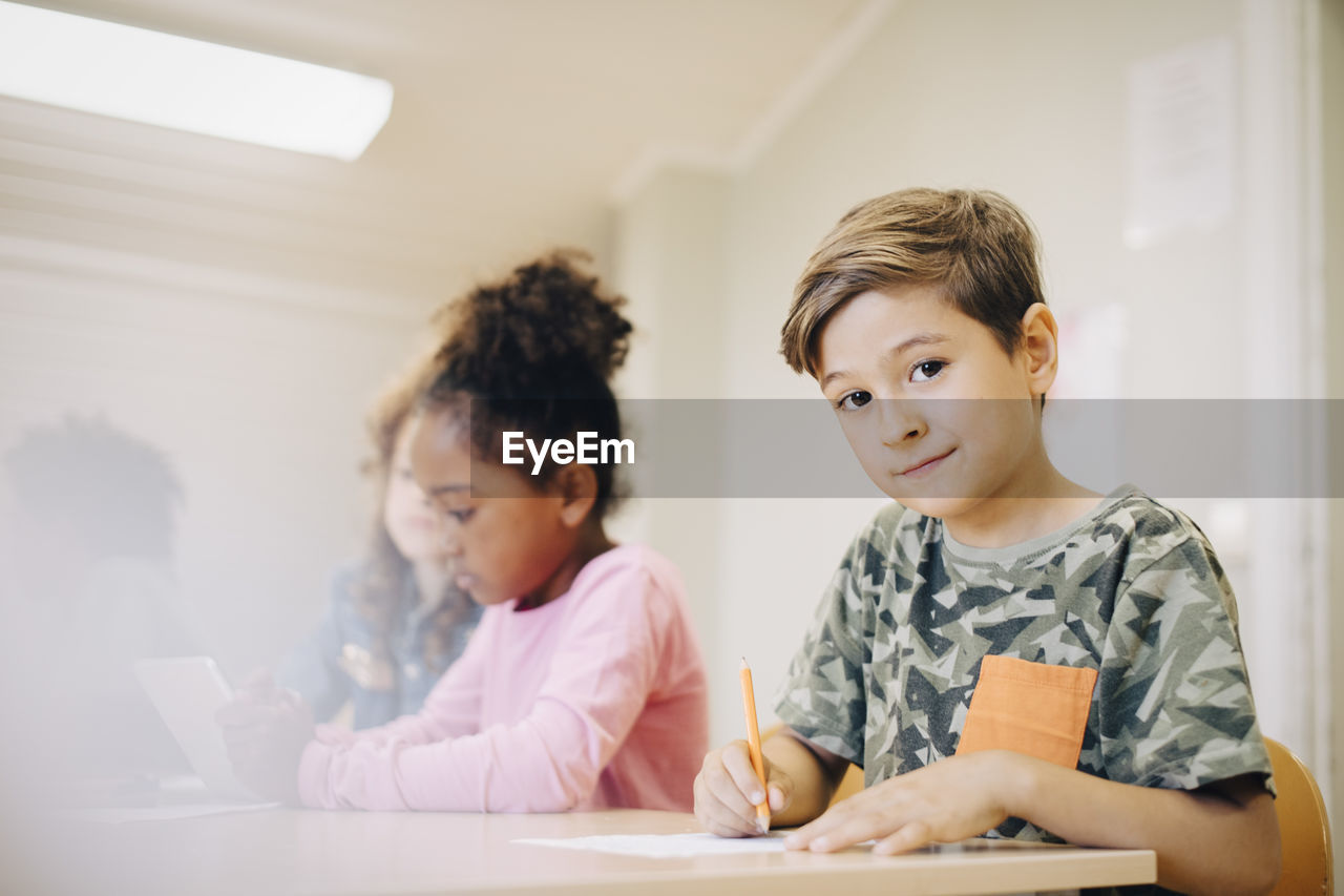 Portrait of boy sitting with friend writing on paper at desk in classroom