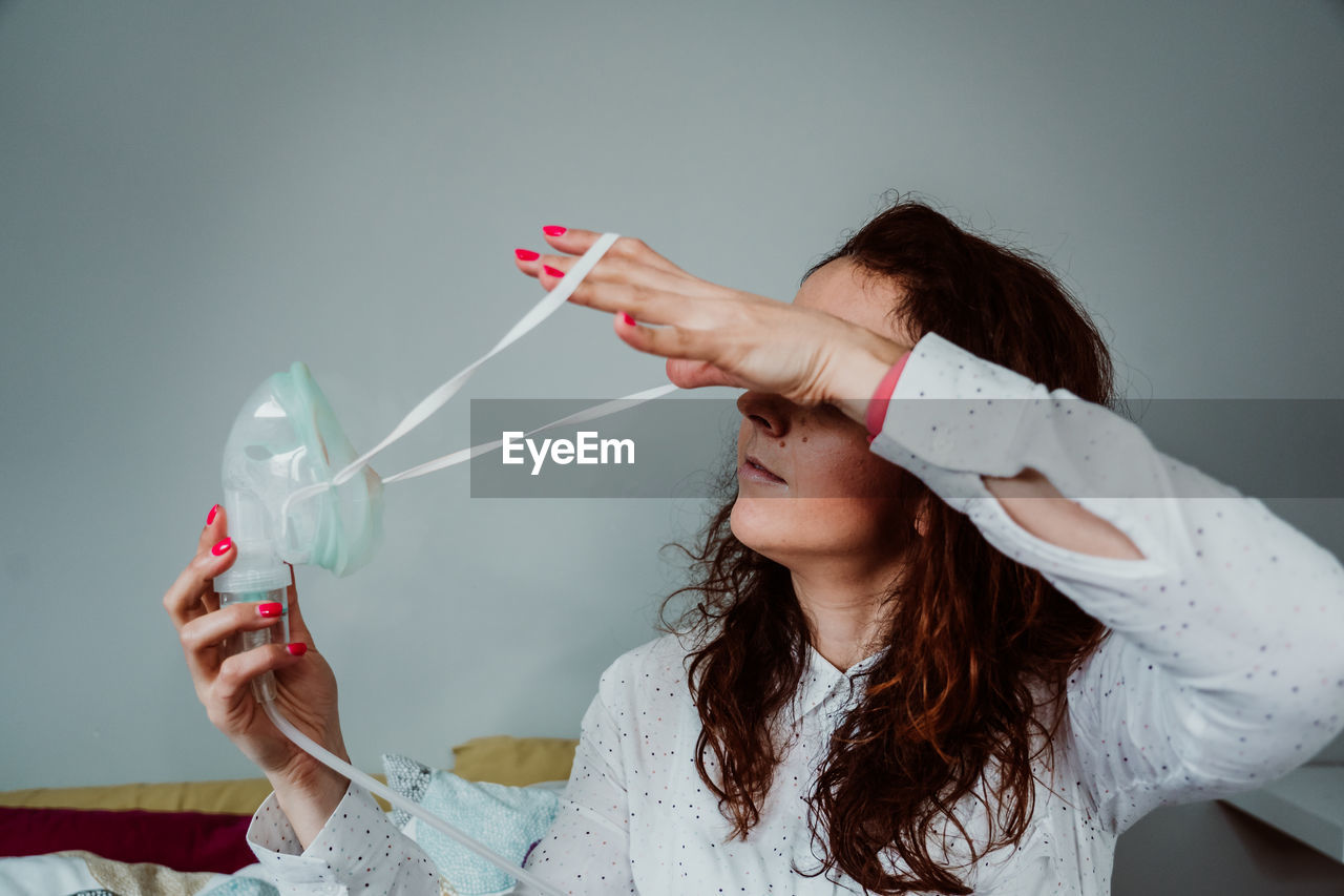 Woman wearing oxygen mask while sitting on bed against wall in hospital