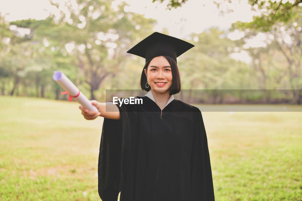 Portrait of young woman in graduation gown holding certificate while standing at park