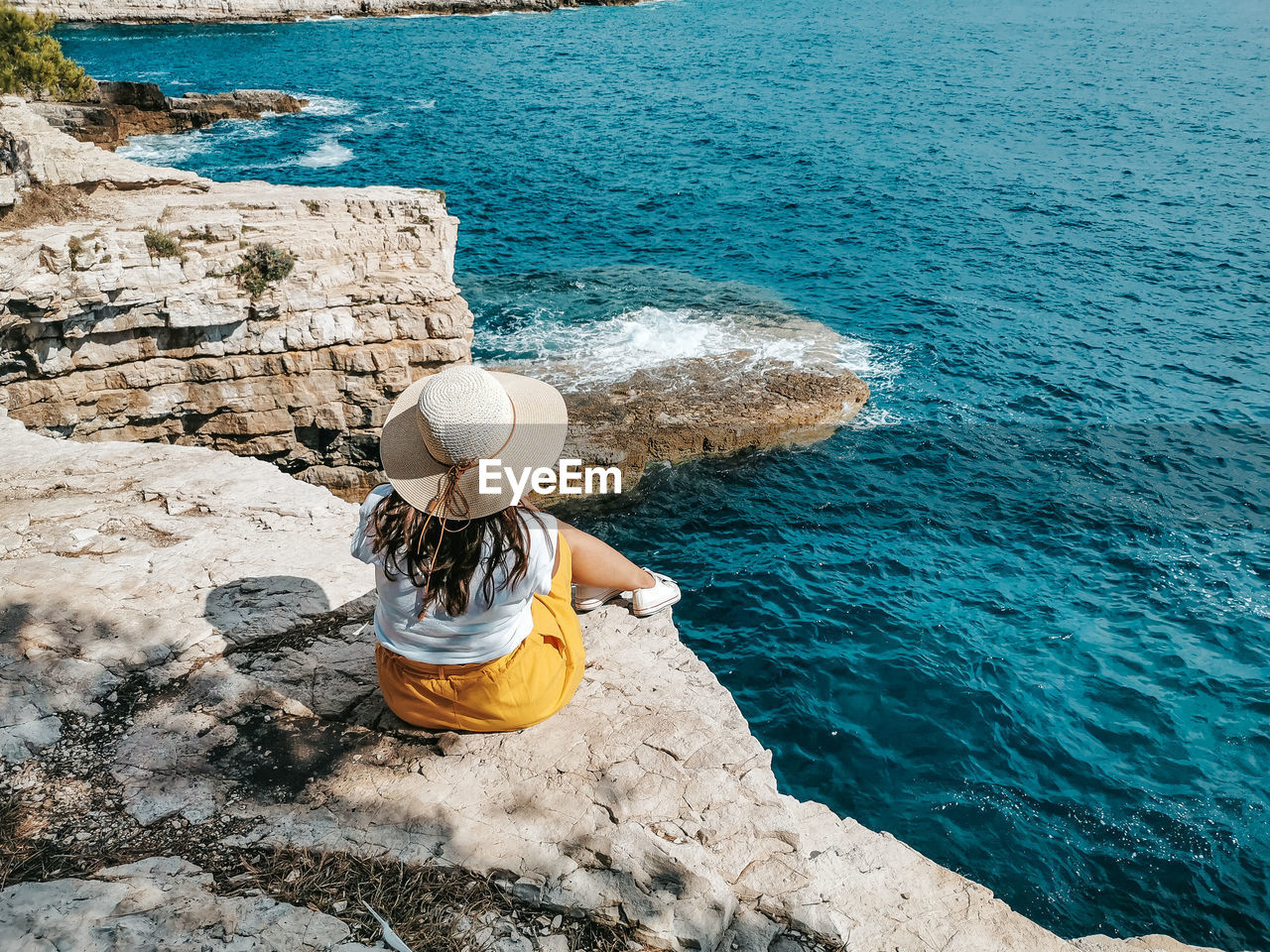 Young woman in yellow skirt sitting on edge of cliff above sea.