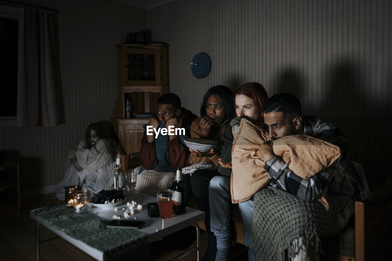 Frightened multiracial friends watching scary movie while ghost sitting in corner at cabin