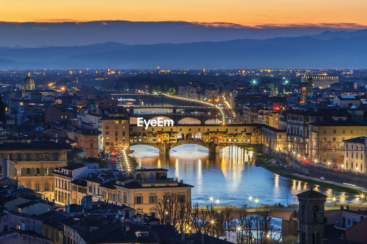 HIGH ANGLE VIEW OF ILLUMINATED CITY BY RIVER AT DUSK