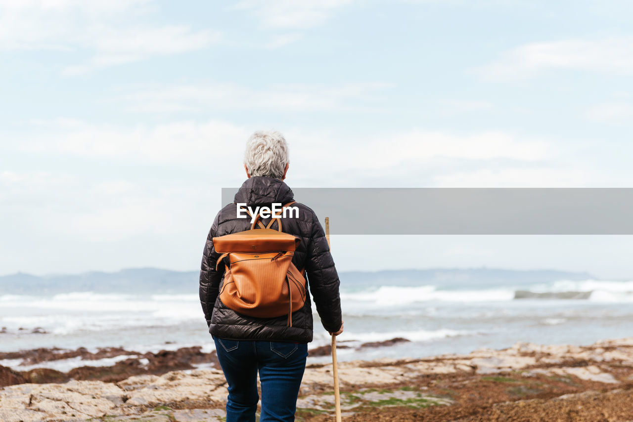 Back view of unrecognizable elderly female backpacker with trekking pole strolling on boulders against stormy ocean under cloudy sky