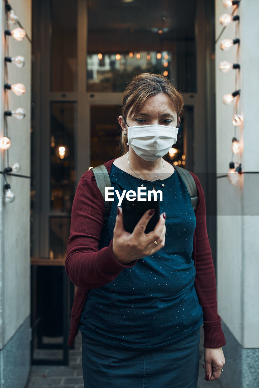 Young woman talking on phone standing in a front of restaurant downtown wearing the face mask