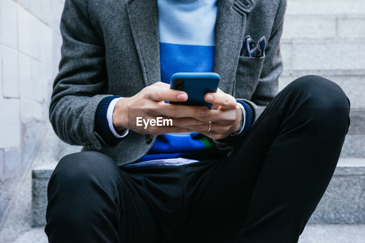 Crop faceless person in smart casual style sitting on stairs in city and messaging on mobile phone