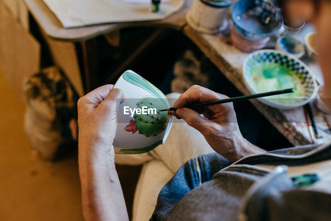 Anonymous focused mature craftswoman with paintbrush painting ceramic bowl while making earthenware in pottery workshop