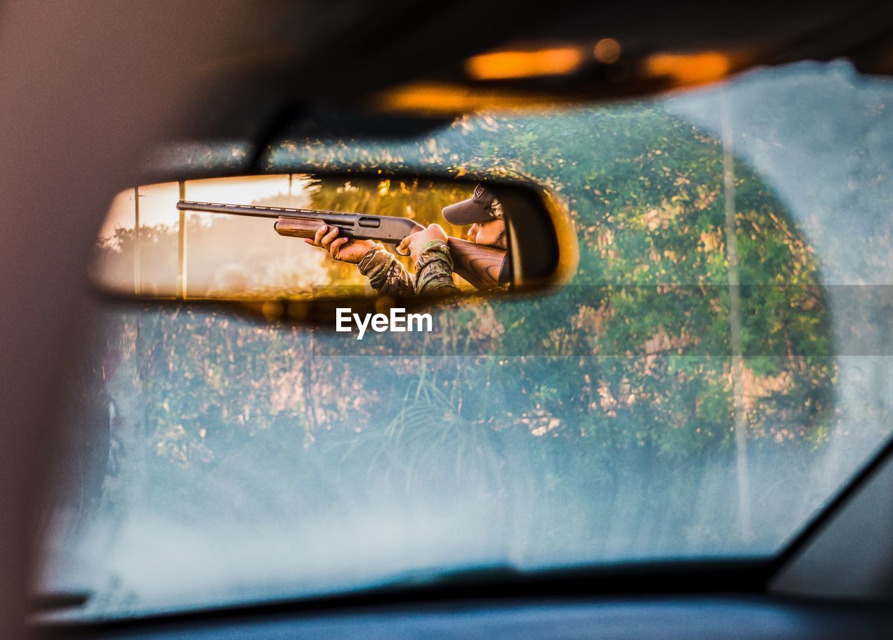 Hunter aiming with shotgun reflecting in rear-view mirror of car