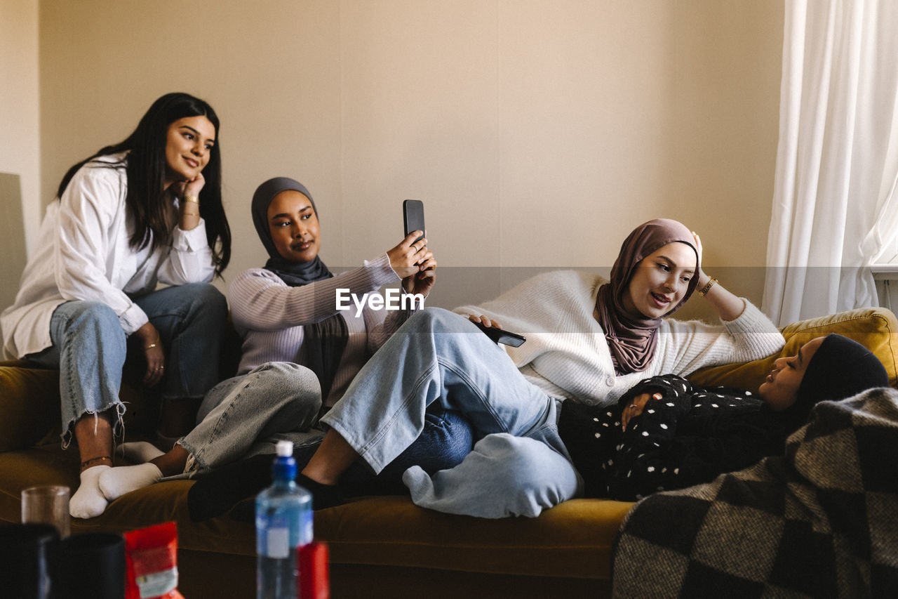 Young women talking with each other by friends taking selfie through mobile phone at home