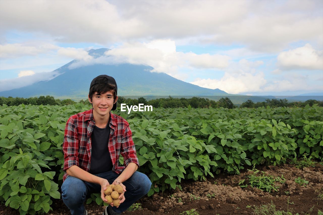 Portrait of smiling teenage boy holding raw potatoes by plants on field against cloudy sky