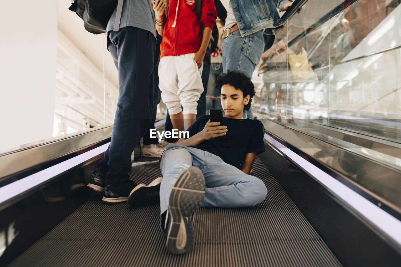 Teenage boy using mobile phone while sitting by friends on moving walkway in shopping mall