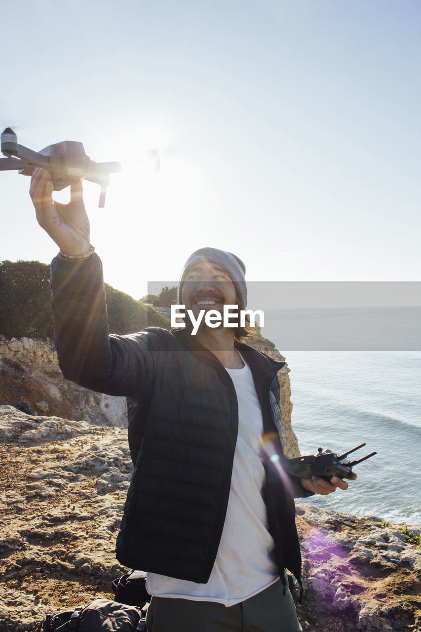 Smiling man flying quadcopter while standing at beach against clear sky during sunny day