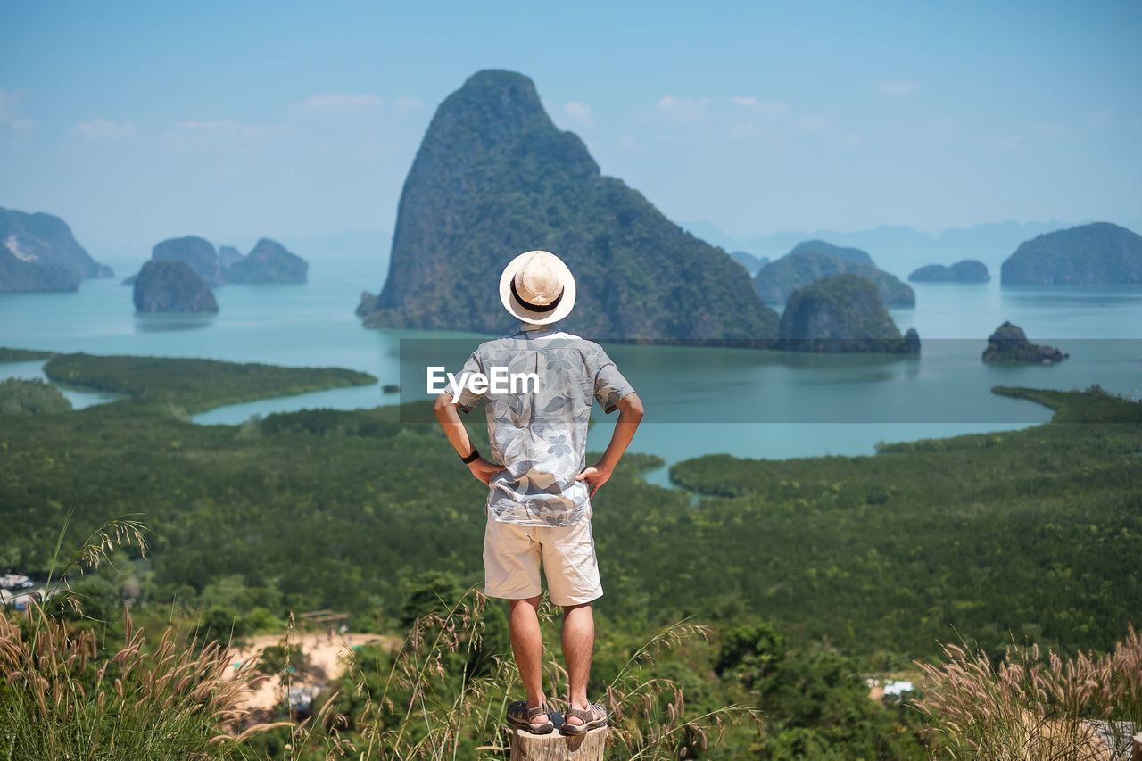 REAR VIEW OF MAN STANDING ON MOUNTAIN AGAINST SKY