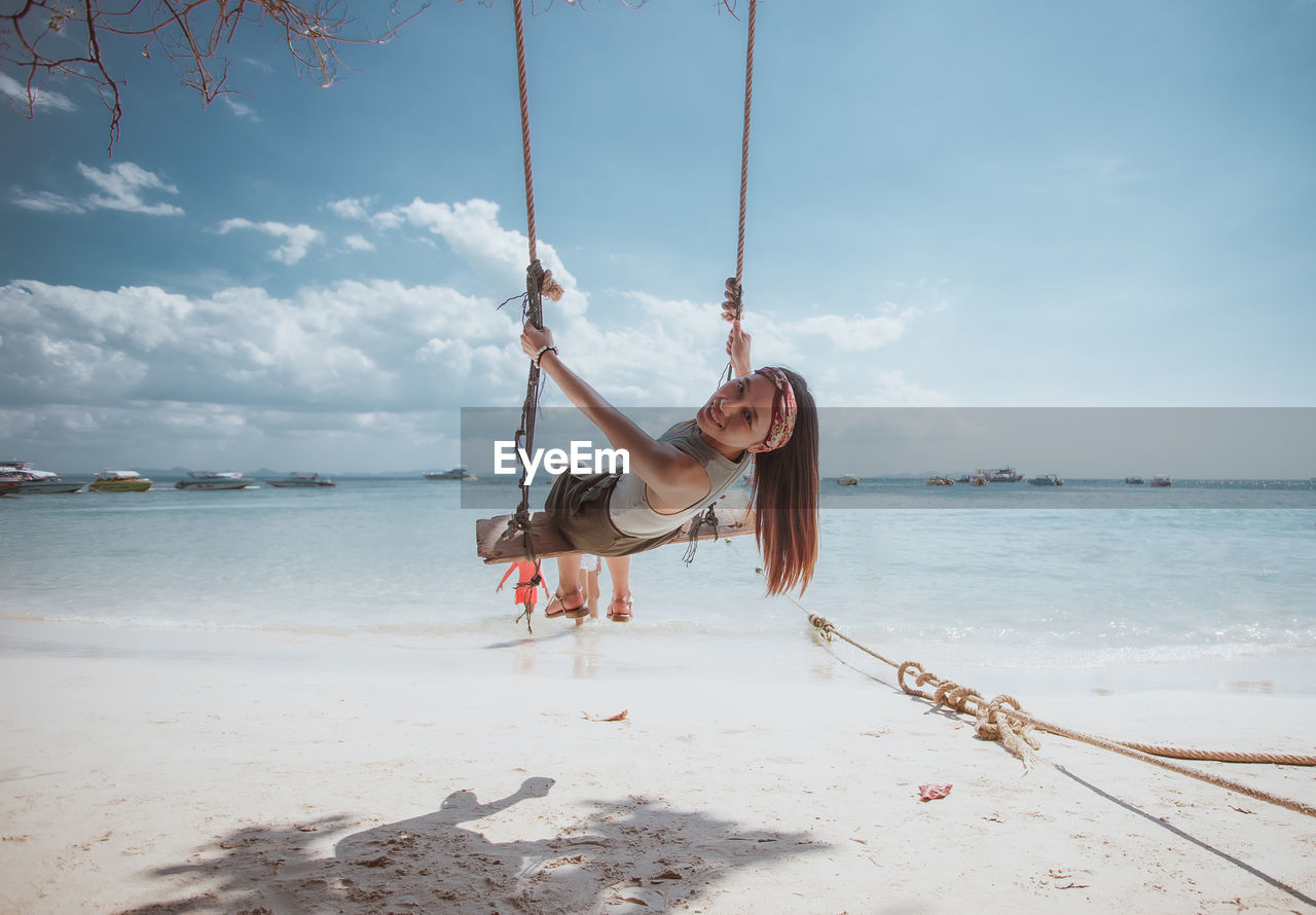 Portrait of smiling young woman sitting on swing at beach against sky