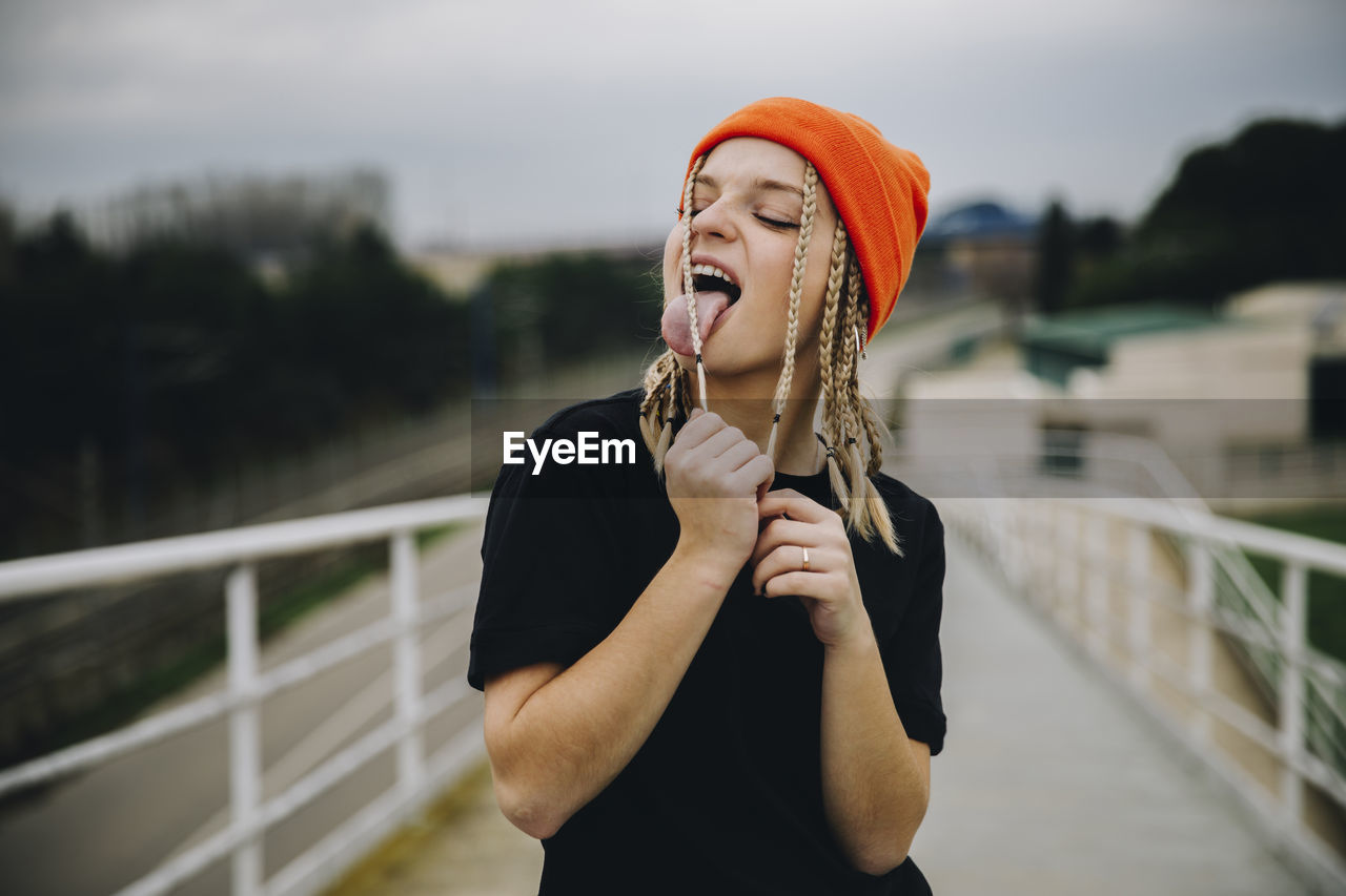 Young woman licking braided hair while standing on footbridge