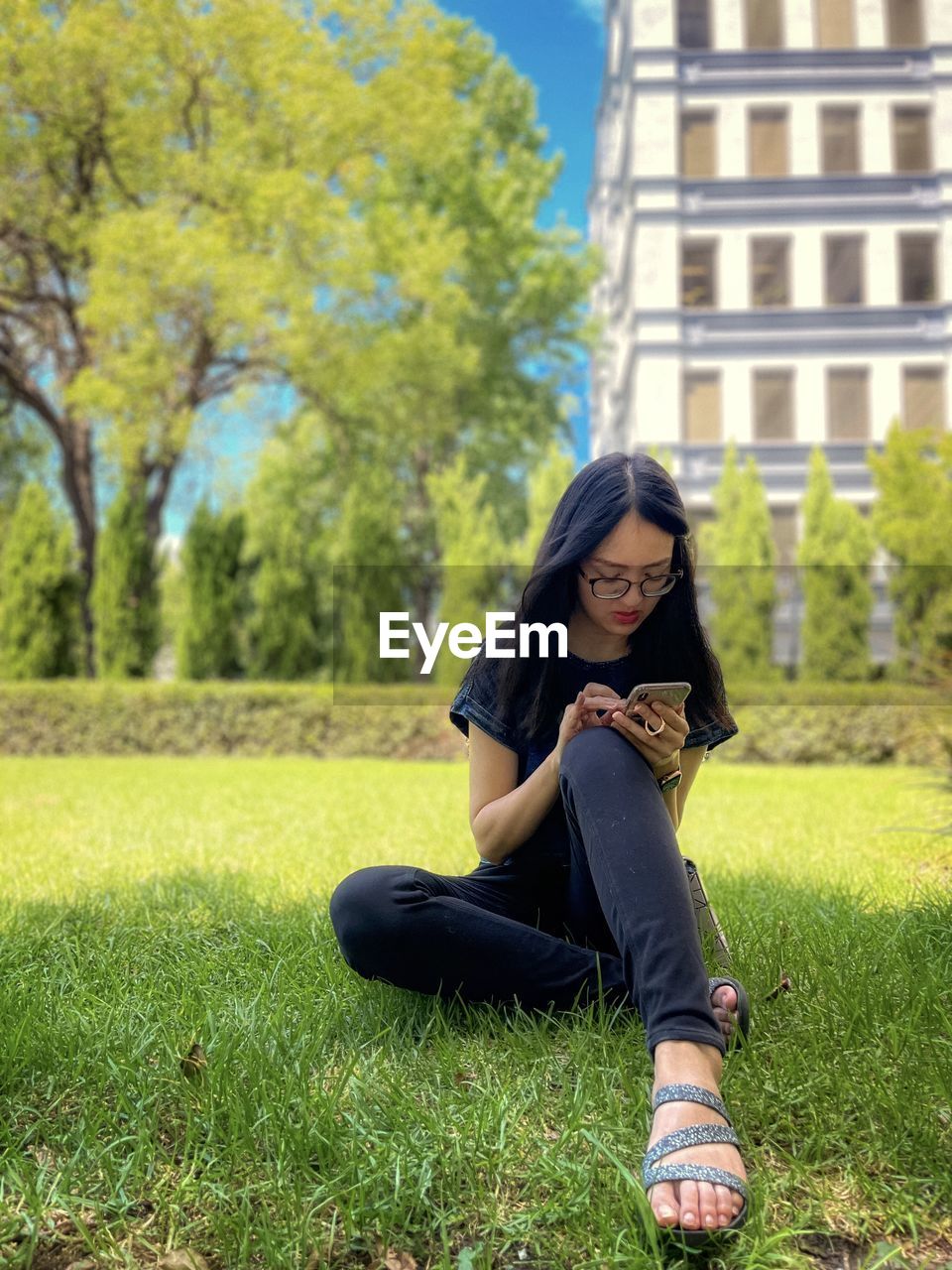 Asian woman sitting on the grass while using smartphone against greenery and building.