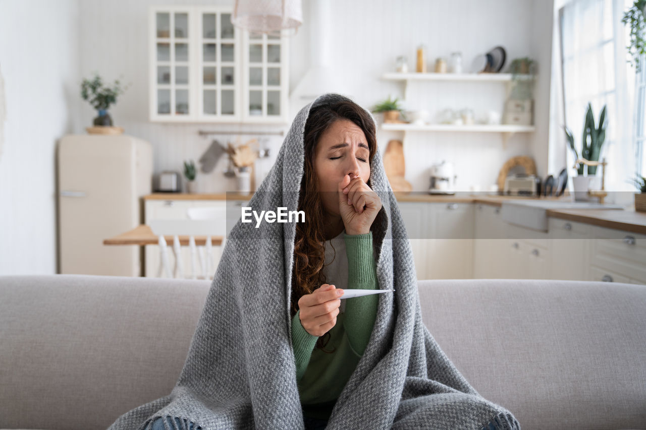 Sick woman coughing, measuring temperature with thermometer, sitting on sofa wrapped in blanket