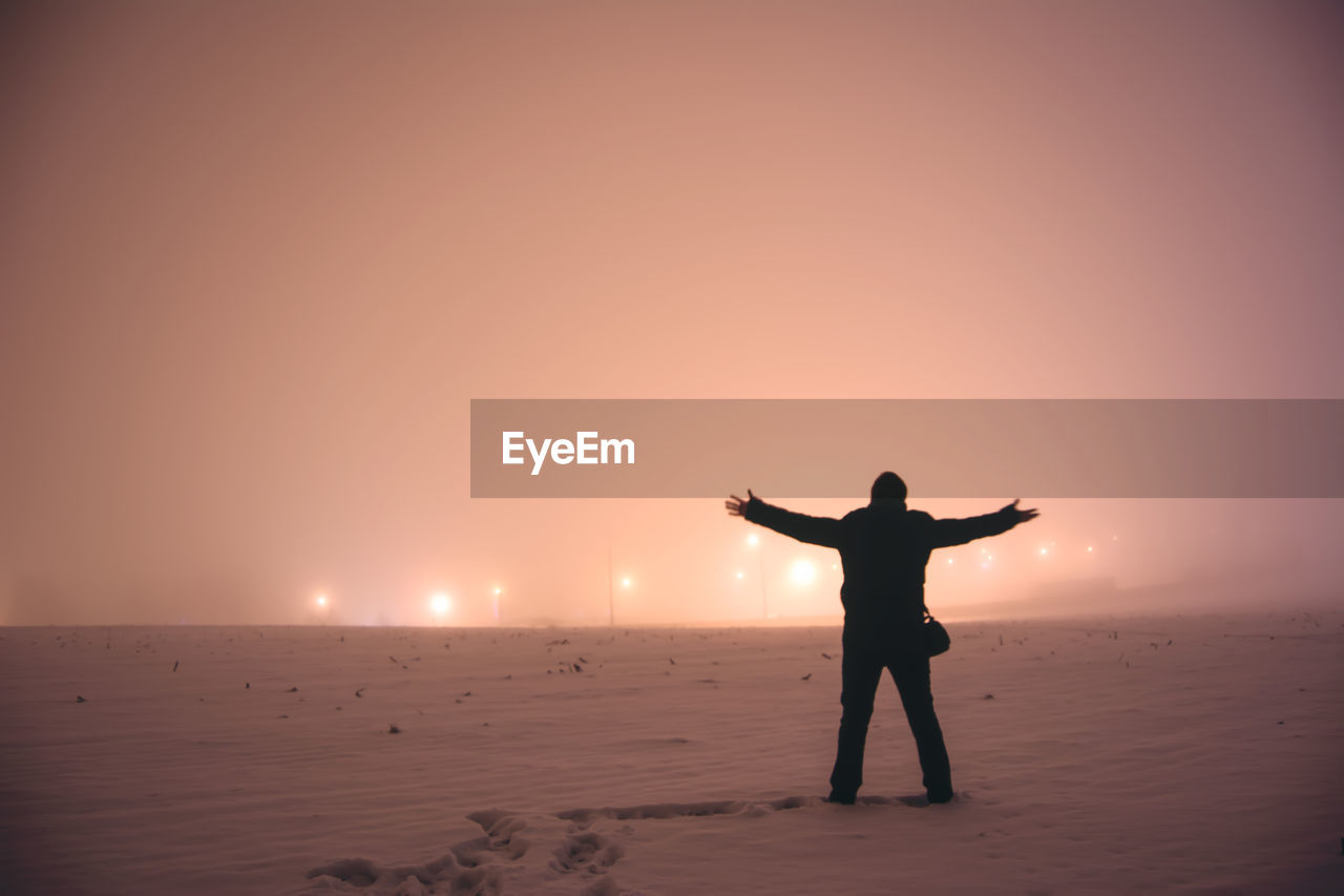 Rear view of man with arms outstretched standing on snow covered field at night