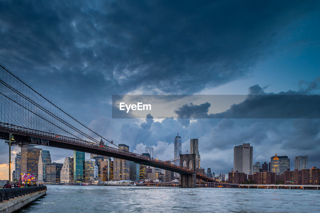 Low angle view of brooklyn bridge over river against cloudy sky in city