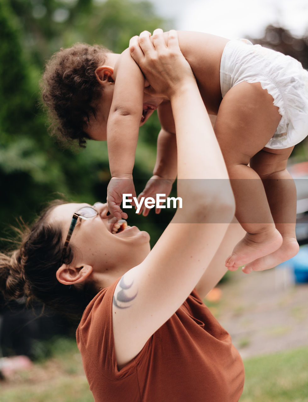 Side view of mother holding baby mid-air standing outdoors