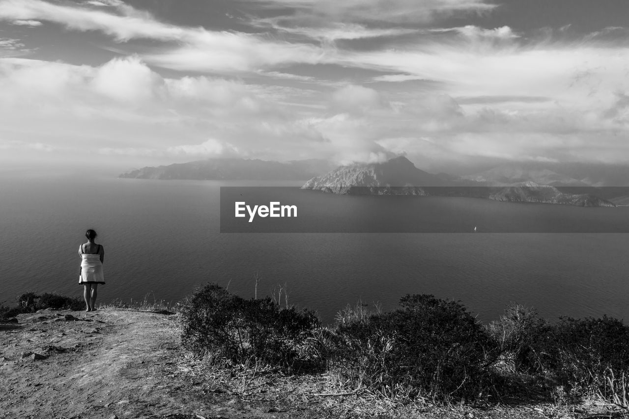 Rear view of woman looking at sea while standing on mountain against cloudy sky