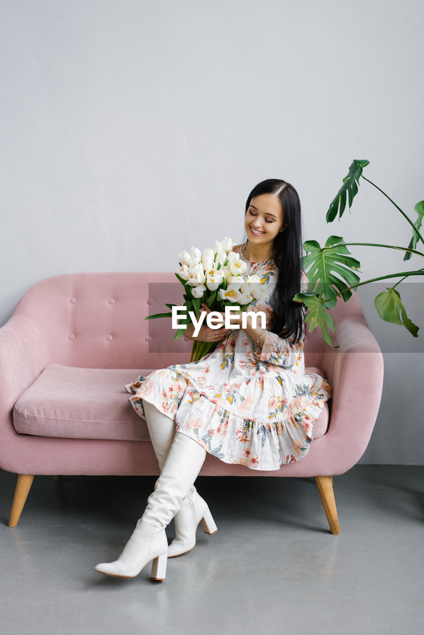 A young pretty happy woman in a dress and white boots sits on a pink sofa with a bouquet