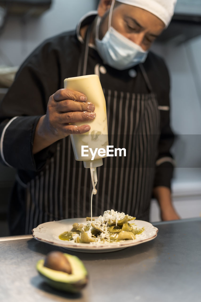 Crop middle aged ethnic male cook pouring white sauce from bottle on delicious pasta with grated cheese in restaurant kitchen