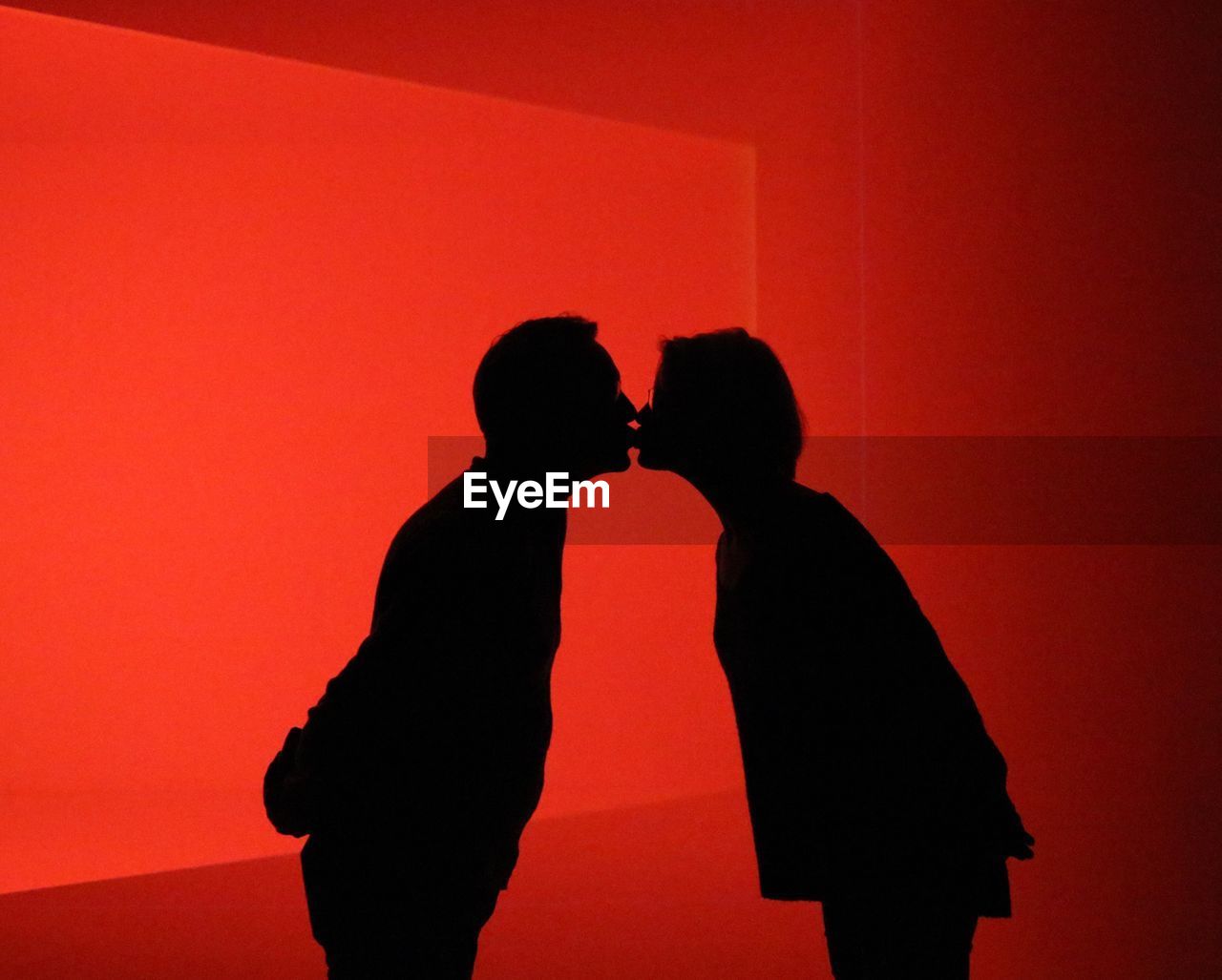Side view of silhouette couple kissing on mouth while standing against red background