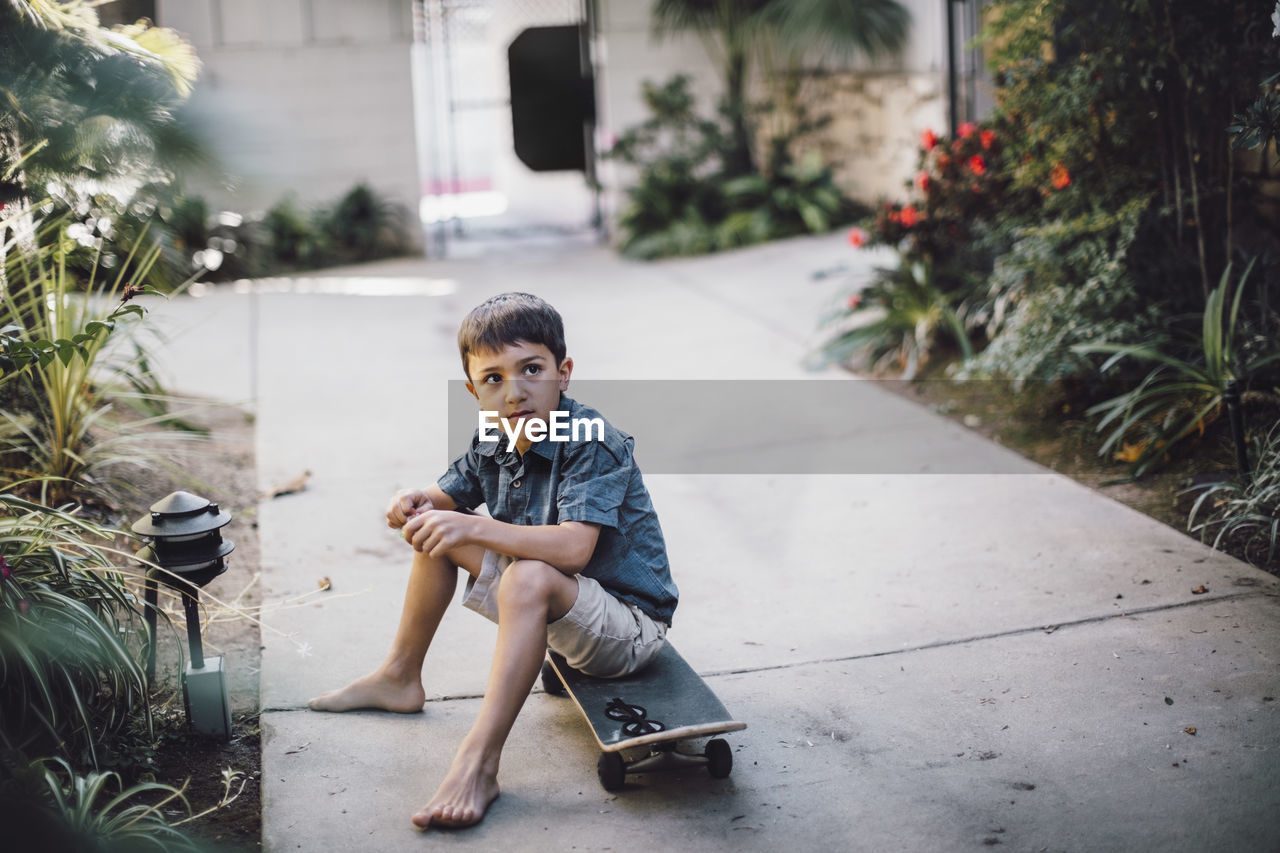 Boy looking away while sitting over skateboard on footpath