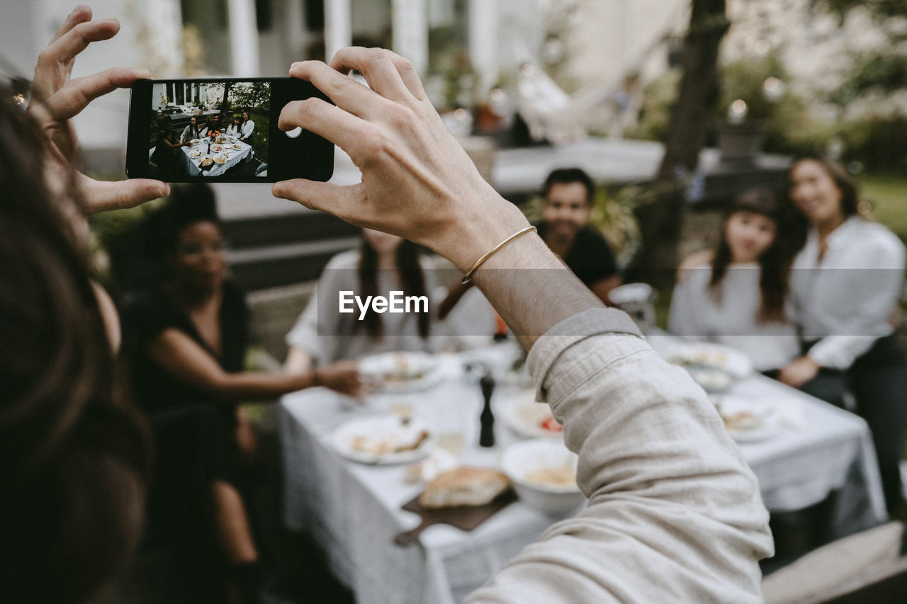 Cropped hand of man photographing through smart phone while friends sitting by table in yard