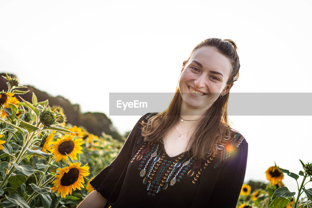 Portrait of smiling young woman standing by sunflowers against sky