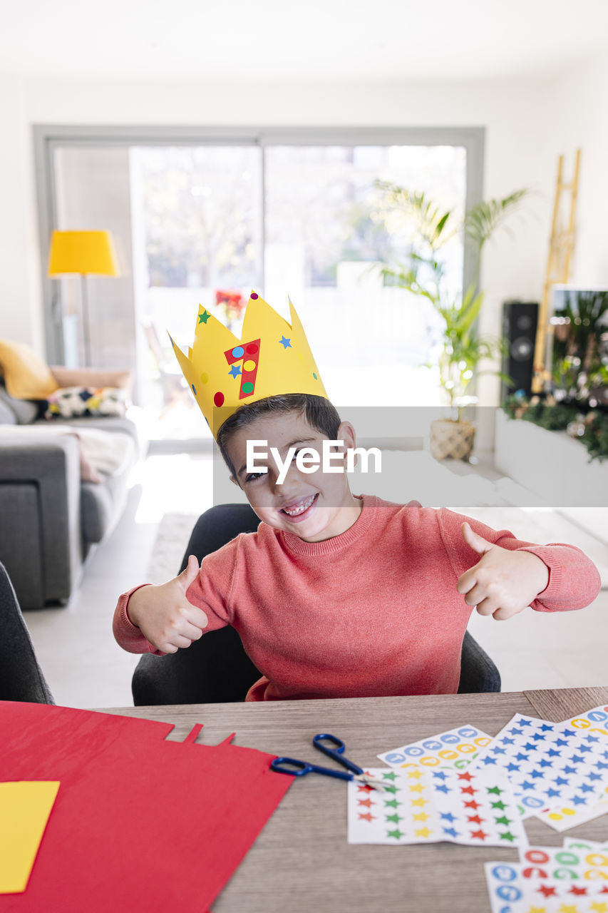 Portrait of boy wearing crown sitting by table at home