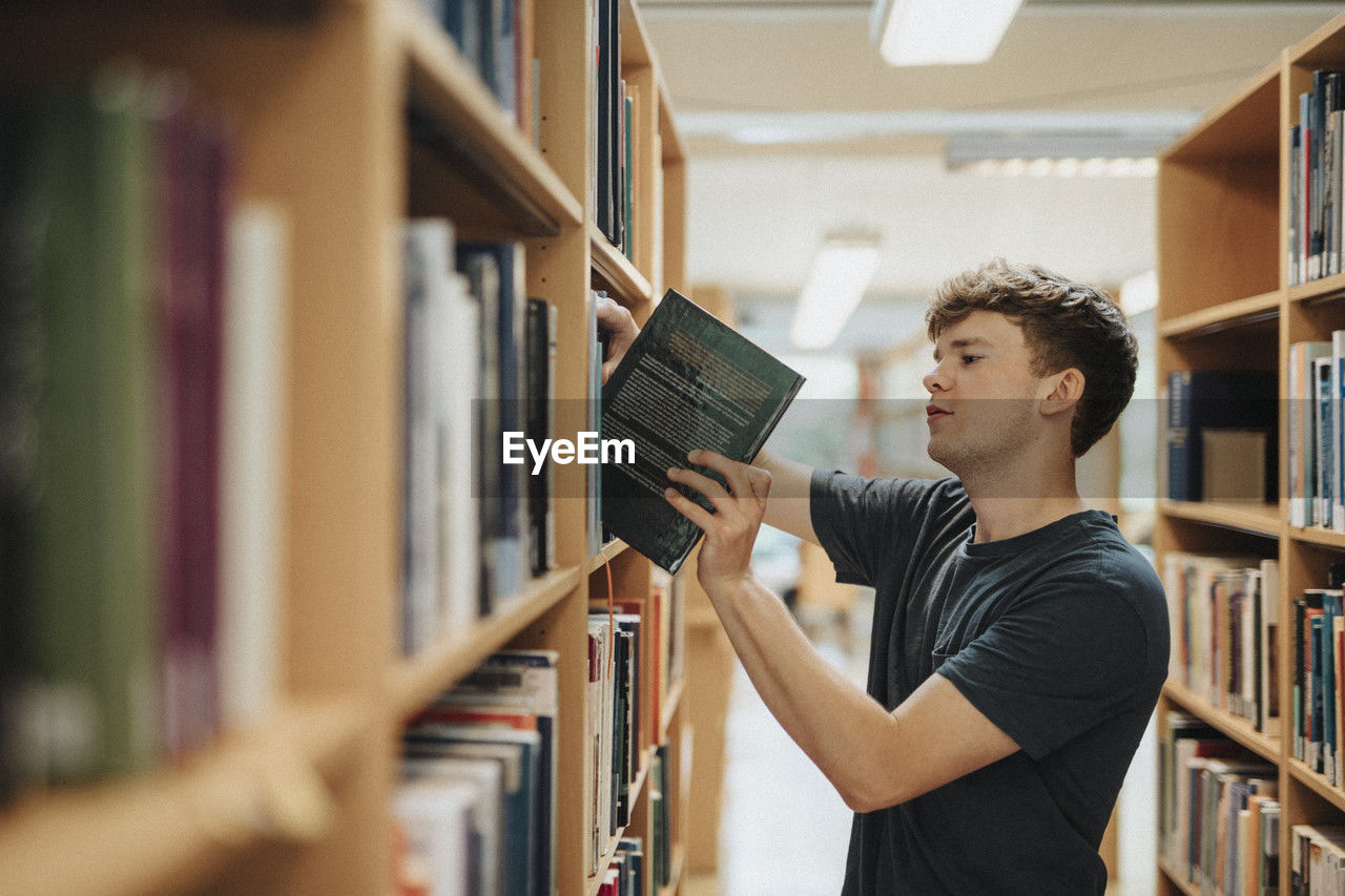 Side view of male student removing book from bookshelf in library at university