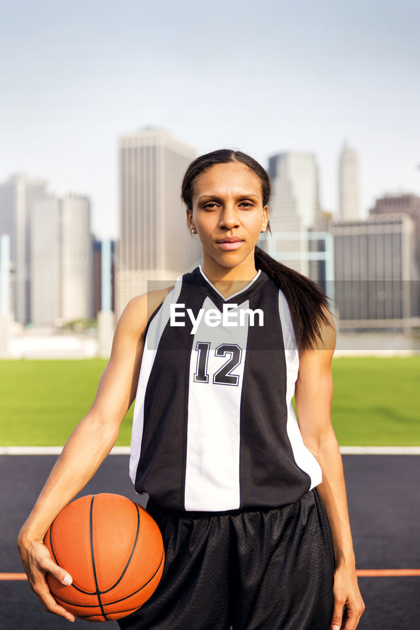 Portrait of woman holding basketball standing in court against buildings