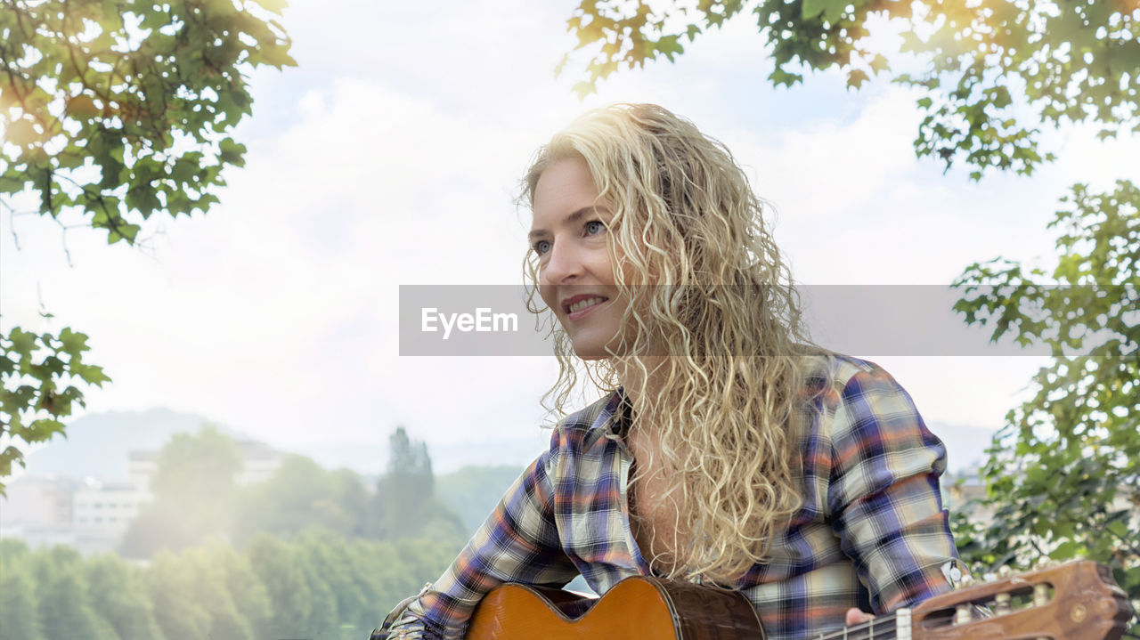 Smiling woman looking away while playing guitar against trees