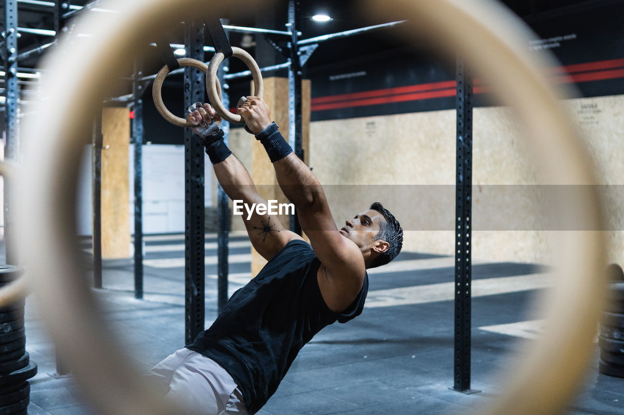 Side view of through blurred sportive equipment of side view strong hispanic male athlete doing exercise with gymnastic rings during training in gym