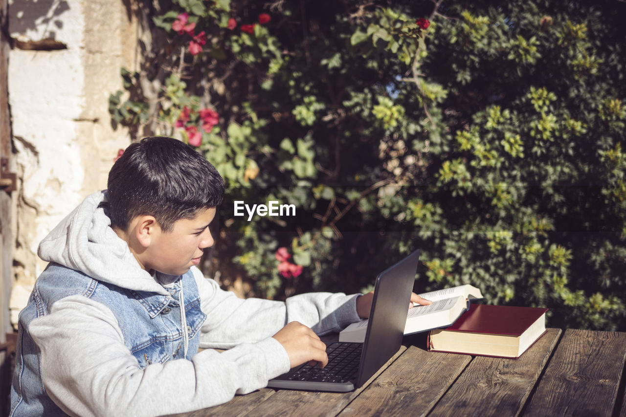 Young latin man reads book in front of laptop, outdoors